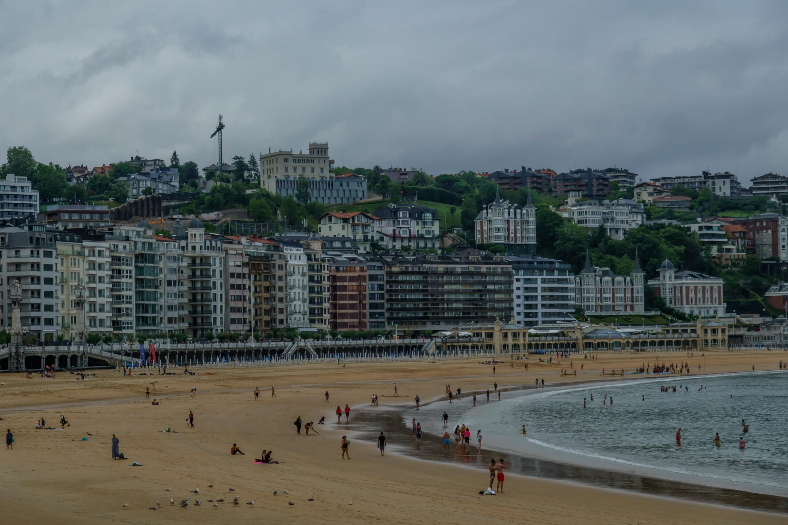 Lush buildings line the sandy beach of San Sebastián, Spain under a cloudy summer sky.