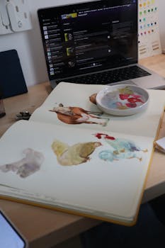 Artistic desk setup with watercolor sketches and paints near a laptop.